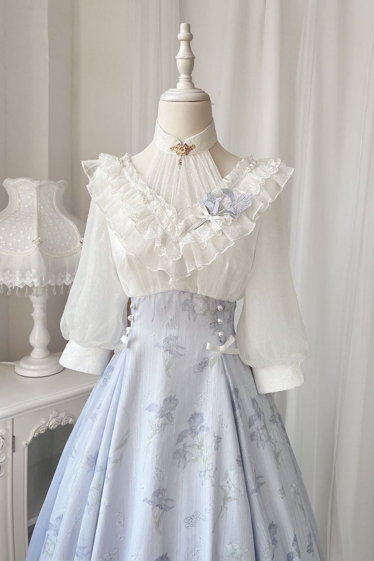 a white and blue dress with a floral print on the skirt. the dress has a high neckline, long sleeves, and a ruffled collar. it is displayed on a mannequin in front of a white curtain.