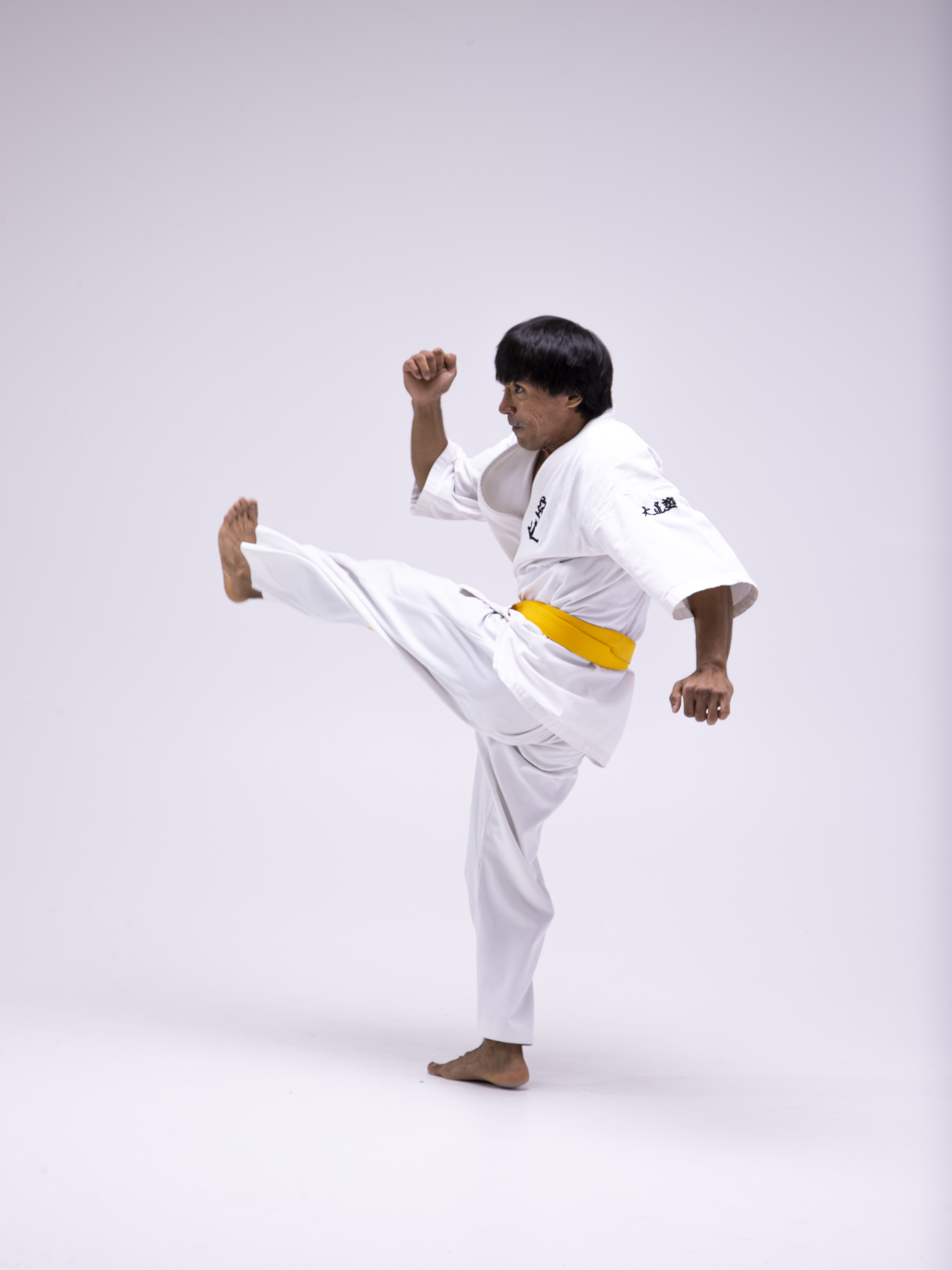 a man wearing a white karate uniform with a yellow belt is performing a high kick. he is barefoot and has his fists clenched. the background is a plain white.