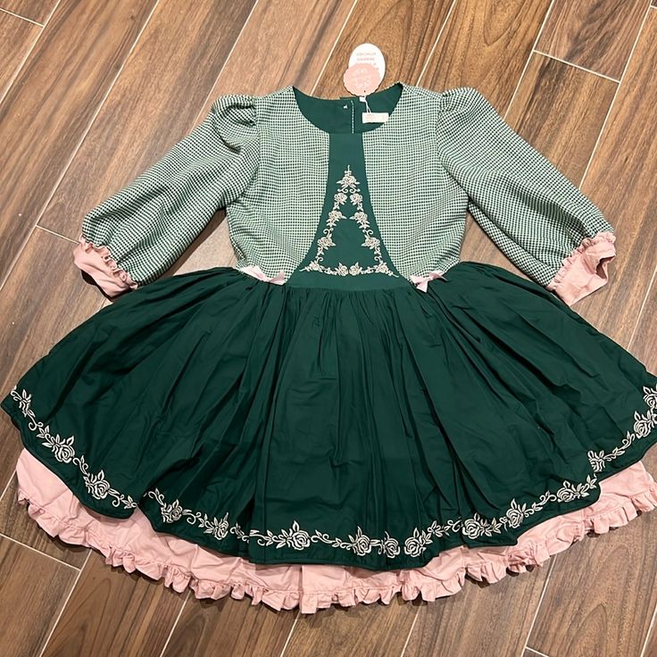 a girl's green and pink dress is laid out on a wooden floor. the top of the dress has a green and white plaid pattern with threequarter length sleeves and a rounded neckline. the skirt of the dress is a solid dark green with pink lining and floral embroidery around the hem. the dress has a vintage or festive style, potentially suitable for christmas or holiday occasions.