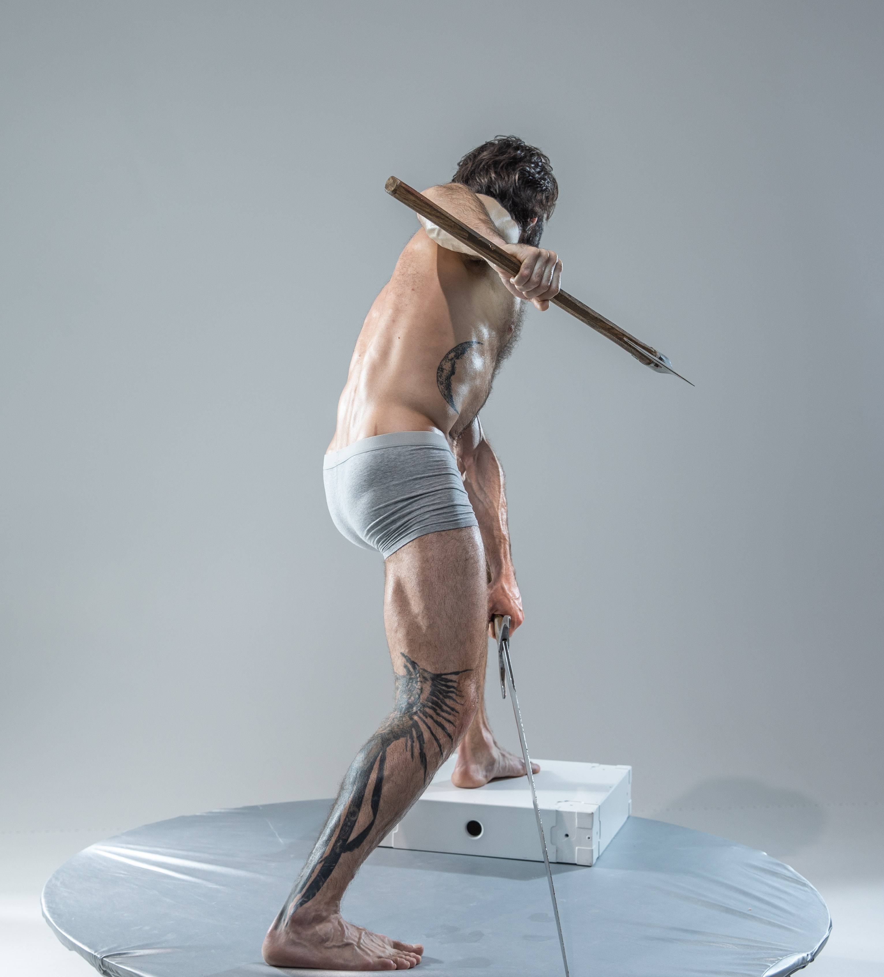 a muscular man with tattoos, wearing only gray underwear, stands on a round platform in a dynamic pose. he holds a wooden axe in his right hand, raised as if to strike, and a sword in his left hand, lowered near the platform. the background is a solid gray, suggesting a studio setting. the lighting is artificial and diffused, casting soft shadows and highlighting the man's physique.