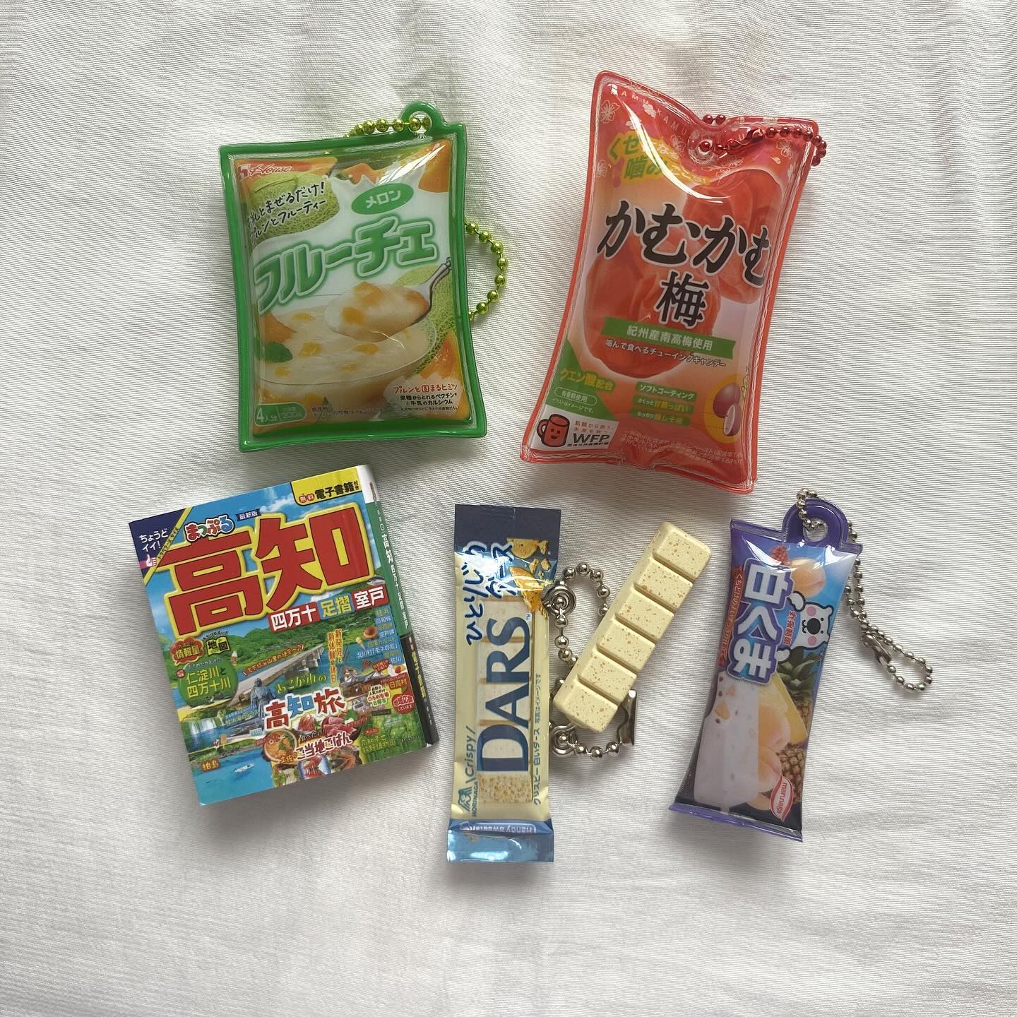 the image shows a flat lay of various japanese candy keychains and a book on a white surface. there are five candy keychains including melon fruit che, ume, dars crispy, white bear and a book titled "kochi".