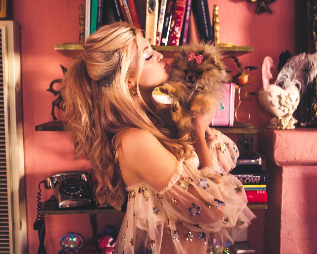 a woman with blonde hair, styled in a halfupdo, is kissing a pomeranian dog held in her arms. the dog has a pink bow on its head. she's wearing a frilly, tulle blouse decorated with stars. they are in a room with pink walls and shelves filled with books, a vintage telephone, a heartshaped swan statue, and other decorative items. a radiator is visible on the left. the overall style is retro and glam, with a feminine aesthetic.