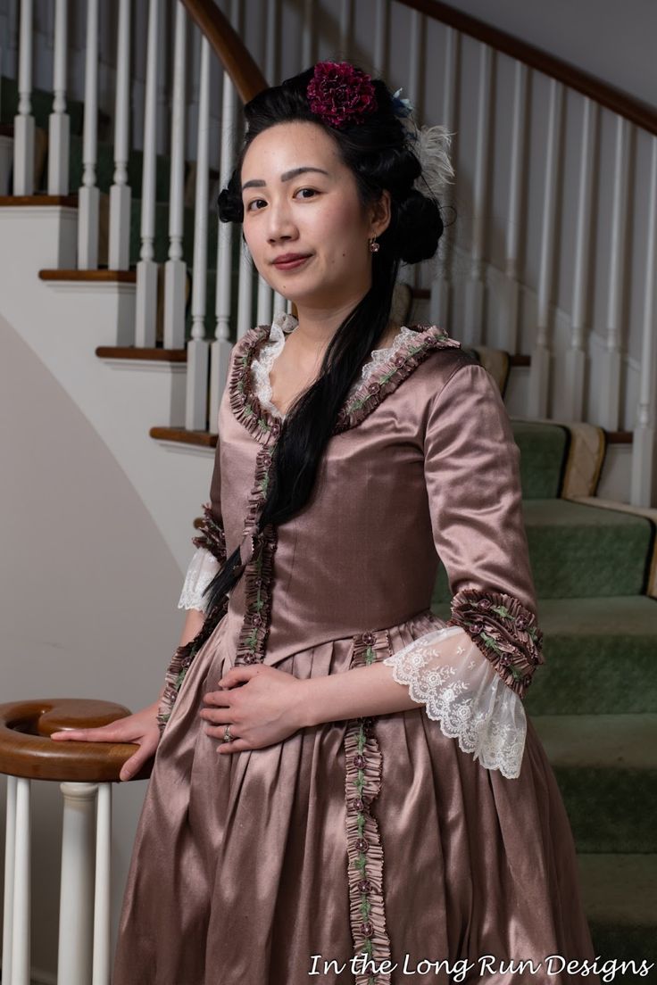 a woman stands in a vintage dress next to a staircase. she is wearing a long, silky brown dress with ruffled edges and white lace details on the sleeves and collar. her black hair is styled in an updo with flowers and a feather. she is standing indoors, leaning slightly on the railing of the staircase behind her.