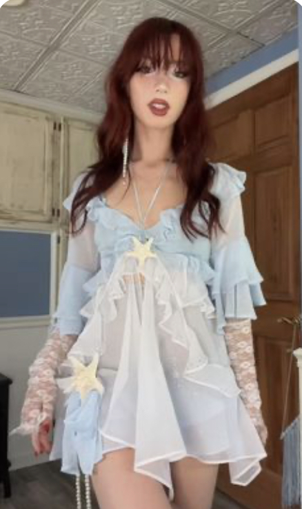 a young woman poses for a selfie indoors. she has long red hair and is wearing a flowy, light blue and white dress. the dress has a ruffled, asymmetrical hemline and is adorned with starfish. she is wearing sheer, lace gloves. she is posing in front of a white door with wood trim. the walls are white and the ceiling is white with blue trim. the room is lit by indoor lighting. the overall aesthetic is whimsical and romantic.