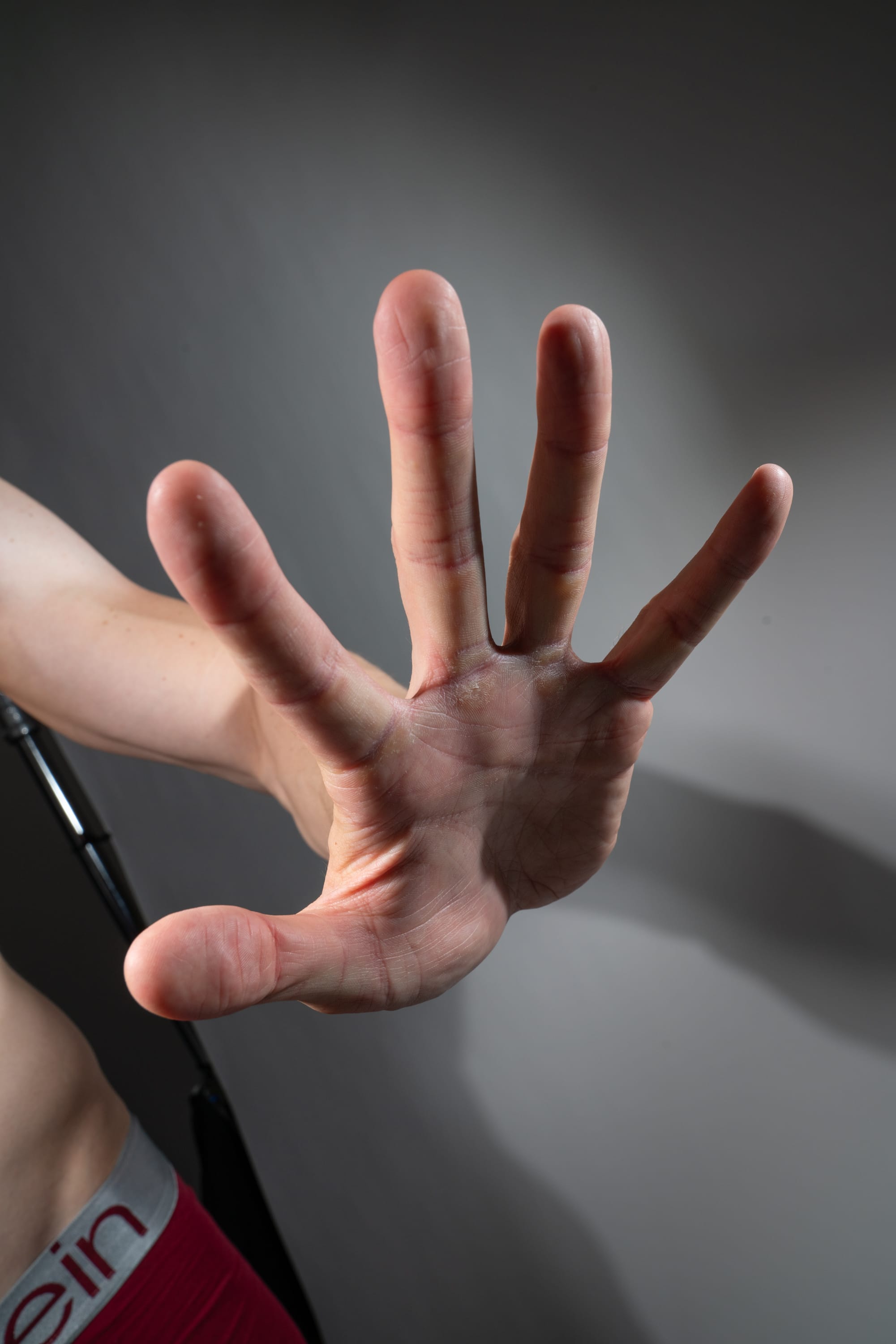 the image shows a closeup of a man's hand held up in a "stop" gesture. the hand is welllit and the details of the skin and lines are clearly visible.  the lighting is soft and diffused, creating a smooth and even illumination. the background is a plain gray wall. the man is wearing red underwear.