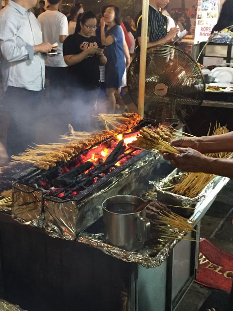 the image shows a bustling street food stall at night, where satay is being grilled over a charcoal fire.  the vendor is actively cooking, tending to the skewers of meat.  smoke billows from the grill, adding to the vibrant atmosphere of the scene. surrounding the stall are people, either waiting for food or enjoying other activities at the market.  the image is welllit, capturing the energy and essence of a busy street food setting. there is also a large industrial fan in the background to help circulate the air and deal with smoke.  a metal cup with what appears to be a marinade or sauce sits on the cart, ready for use.