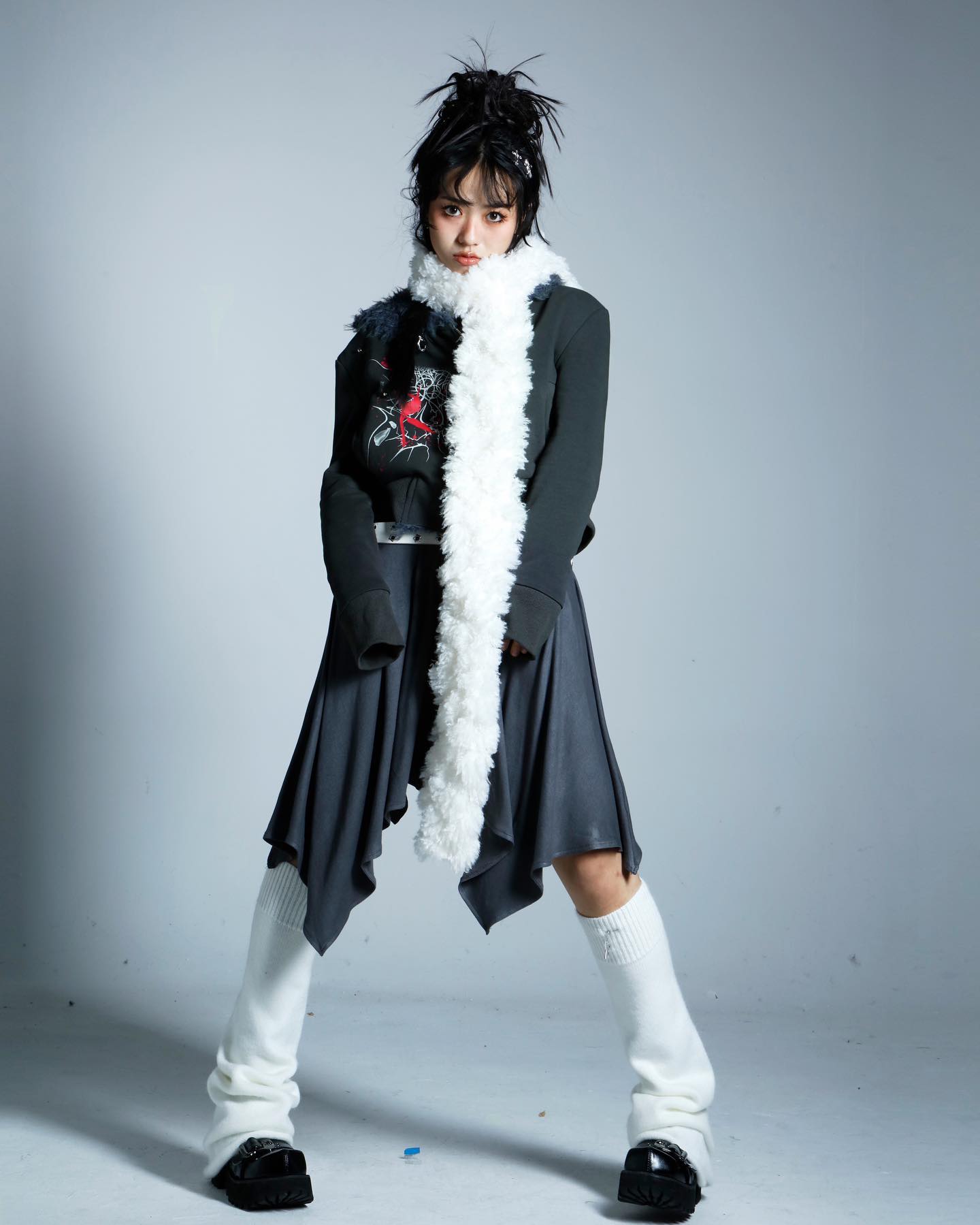 a fullbody studio shot of a young asian woman modeling an edgy, alternative outfit. she's wearing a dark grey asymmetrical skirt and jacket combo over a graphic black and red shirt.  a long, fluffy white scarf is draped around her neck, and she sports white leg warmers and black chunky boots. her hair is styled in a spiky, punkinspired updo. the background is a simple light grey, allowing the outfit and model to be the focus. the overall aesthetic is dark, stylish, and slightly rebellious.