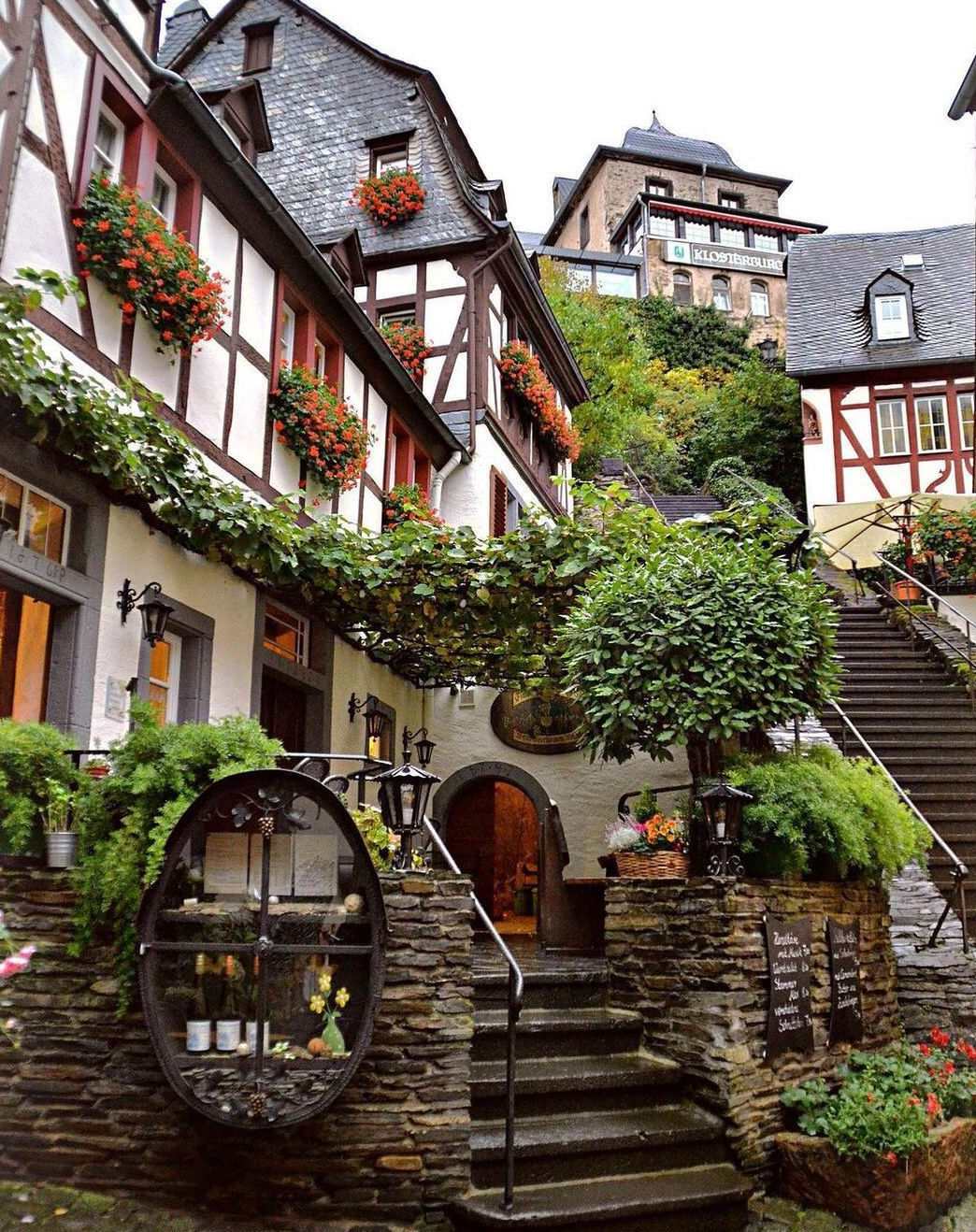 this image showcases a charming european town with traditional buildings adorned with flowers and vines. a staircase leads up to a restaurant entrance, decorated with stone accents, lanterns, and a unique display window. the overall scene evokes a sense of quaintness and oldworld charm.