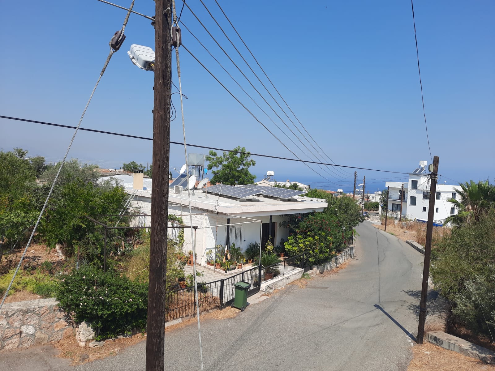 an outdoor daytime shot shows a street and houses, utility poles and power lines, as well as a green trash can and some plants. there is a road going from the bottom left towards the middle right. a house with solar panels on the roof is on the left. there is a second white house further up the street on the right. utility poles line the street. the sky is clear and blue.