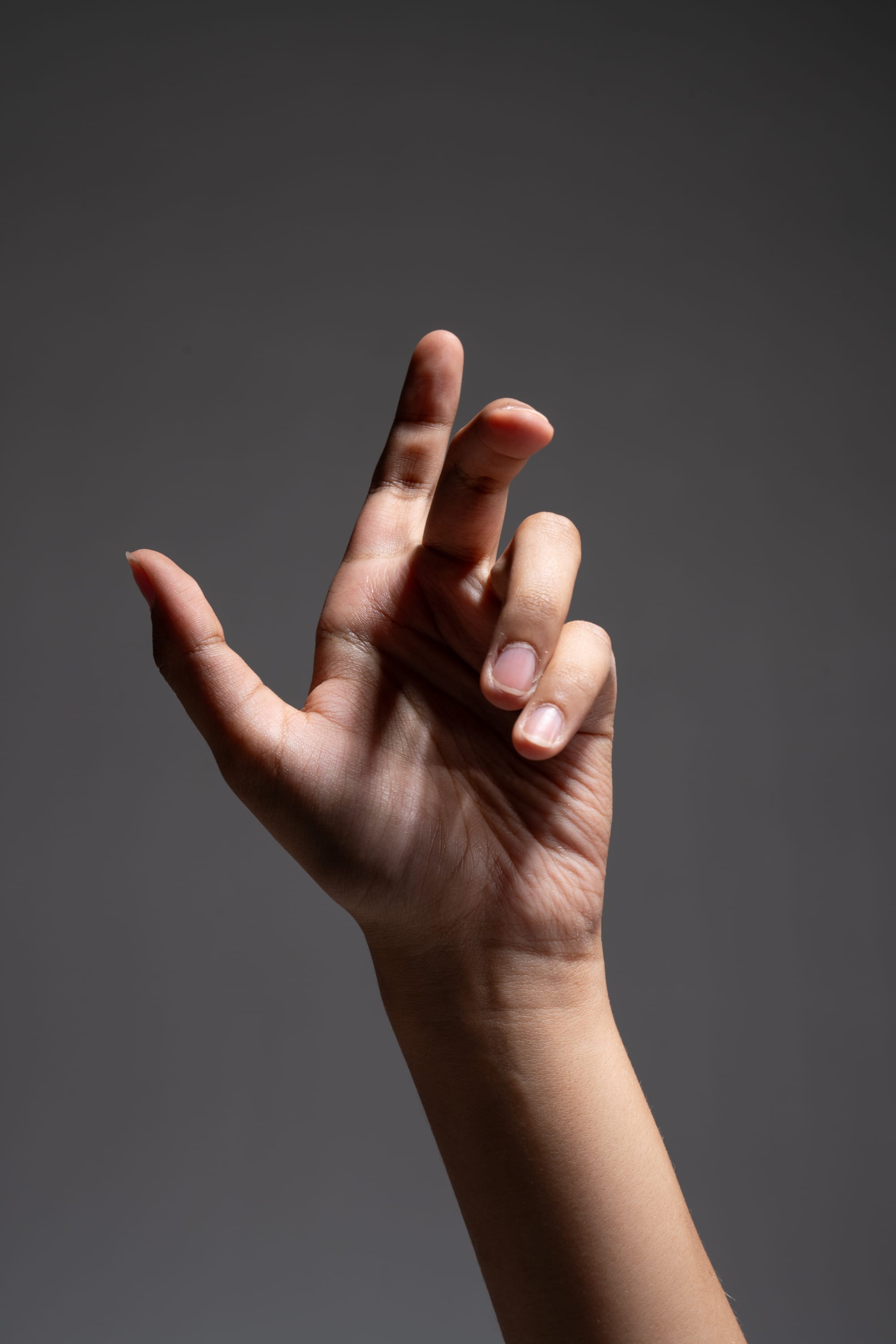 a woman's hand is elegantly posed against a dark grey background. the hand is lit by a spotlight, creating dramatic shadows and highlighting the delicate details of the fingers and skin. the perspective is from the bottom up, making the hand appear larger and emphasizing its graceful form.