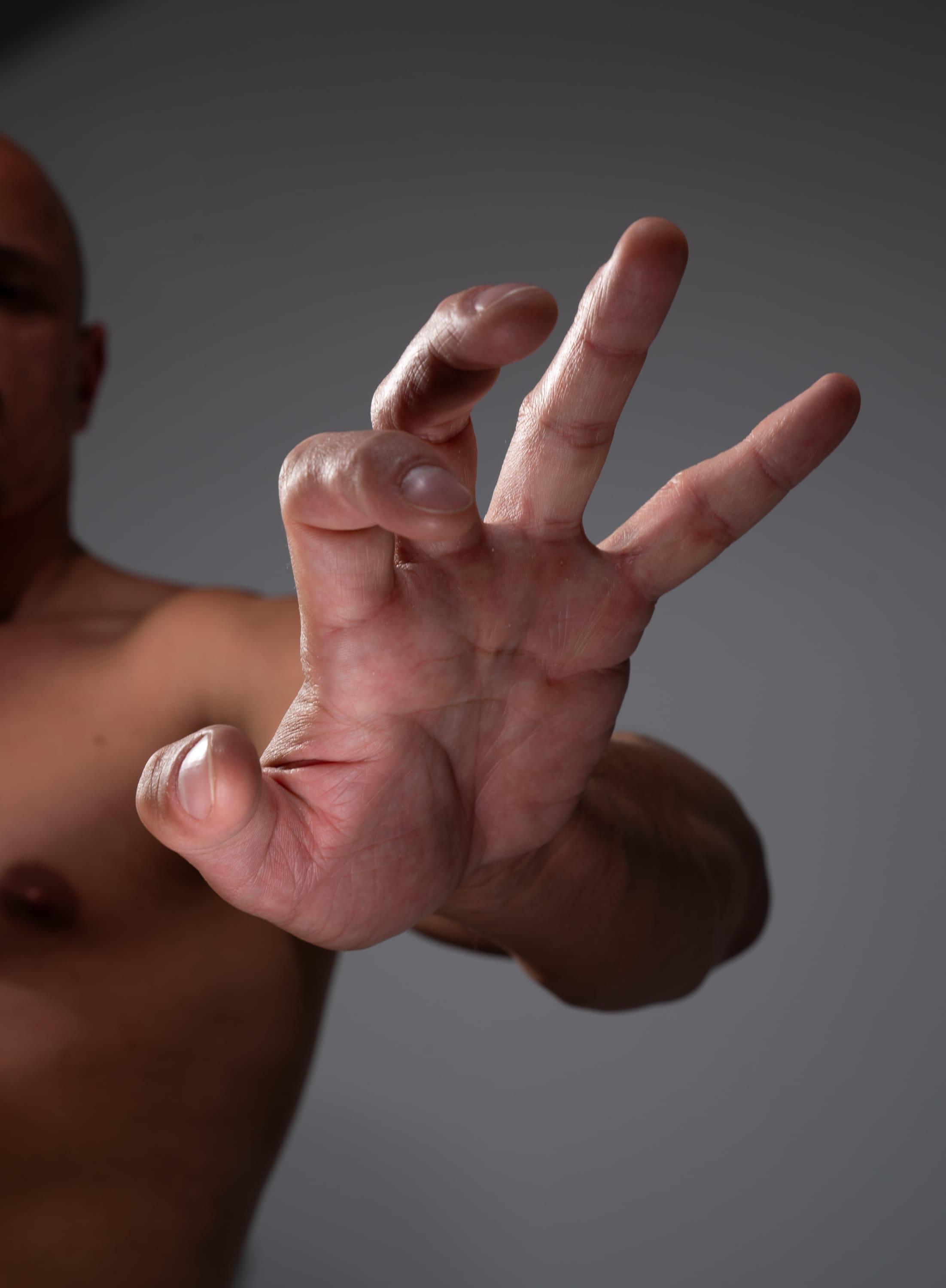 a closeup shot from a low angle of a muscular man with his hand outstretched towards the camera, fingers partially curled. the background is a neutral gray. the lighting is soft and diffused, highlighting the contours of the hand and arm.