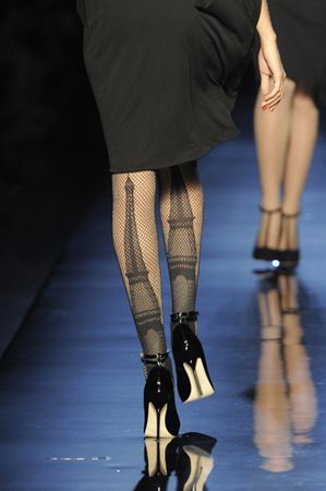 a model is walking on a runway wearing a black dress, tights with eiffel tower prints, and black and white shoes. the runway is reflective, showing the reflection of the model's legs and shoes. there is another model in the background.