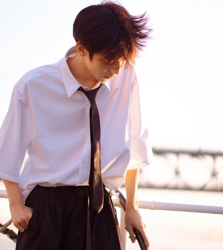 a young asian man wearing a white oversized shirt and black pants stands by a railing on a bridge. he is looking down, and his hair is styled in a windblown look. the background suggests the photo was taken near a body of water at sunset, with warm, natural light illuminating the scene.