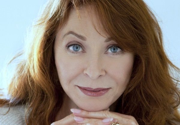 a portrait of a woman with long, auburn hair. she is looking directly at the camera and smiling slightly. she is wearing lipstick and eyeshadow, and she has a gold ring on her finger. she is holding her chin with both hands. the background is blurred.