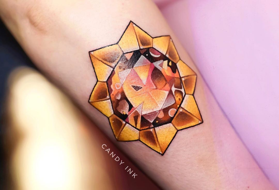 a tattoo of a stylized yellow and orange jewel or crystal on someone's inner forearm. the tattoo has thick black outlines and white highlights to give it dimension. there is text below the tattoo that reads "candy ink".