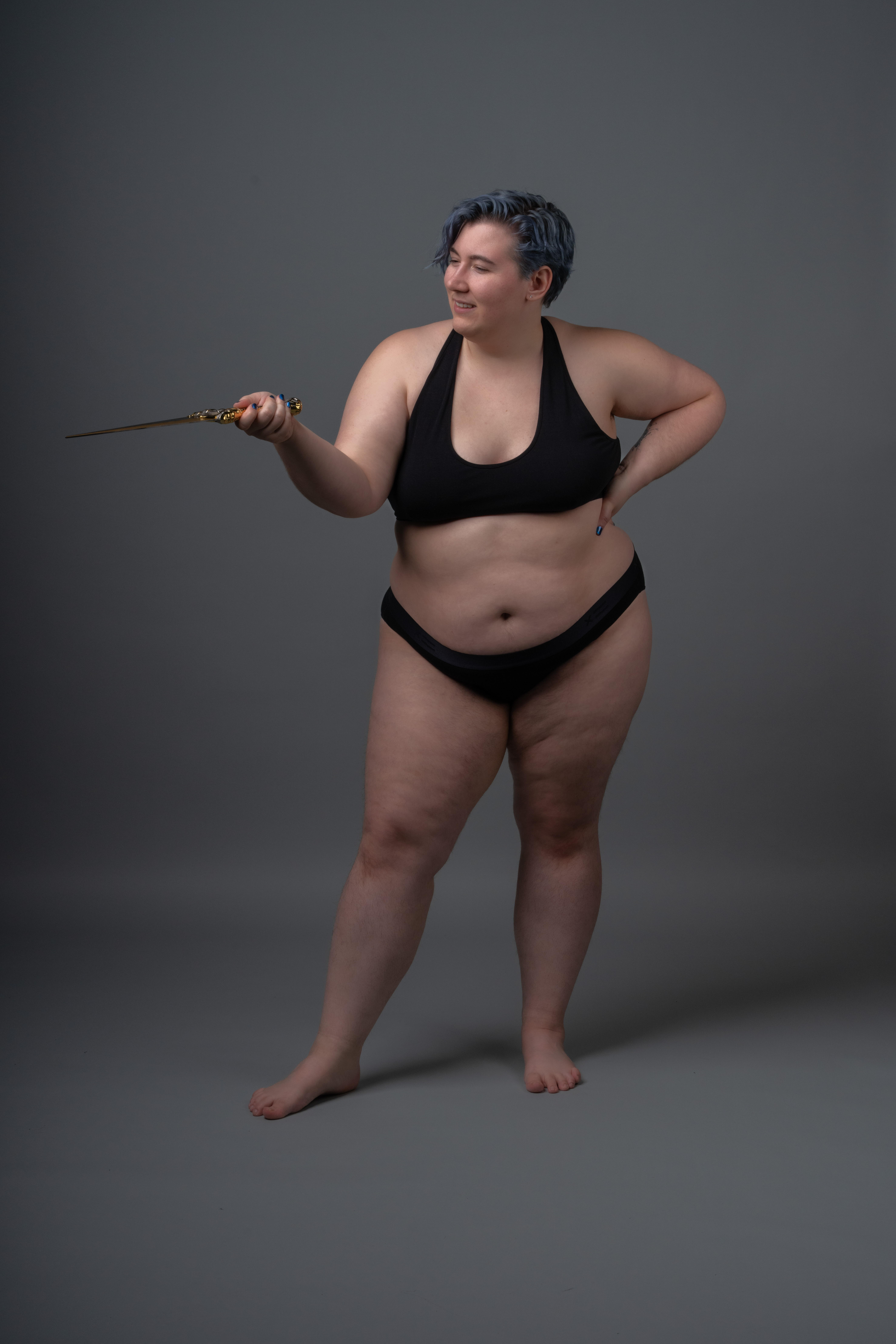 a plussize woman wearing a black bikini is standing indoors against a gray background. she has short blue hair and is smiling. she is holding a knife in her right hand.