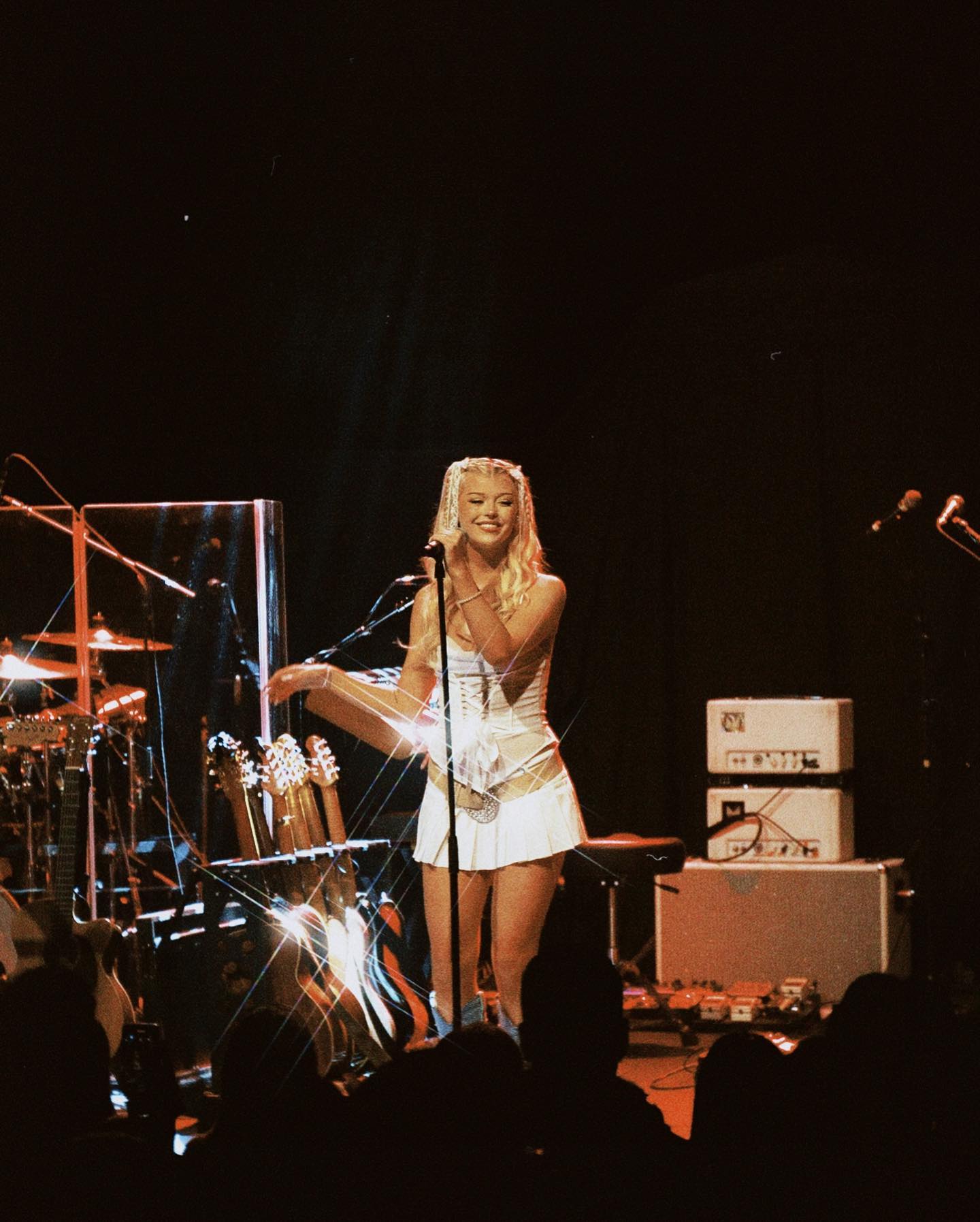 the image shows christina aguilera performing on stage during a concert. she is wearing a white mini skirt and corset outfit. she is singing into a microphone and the stage is lit with bright lights. in the background, there is a guitar rack, amplifiers, and a drum set. an audience is present in the foreground.