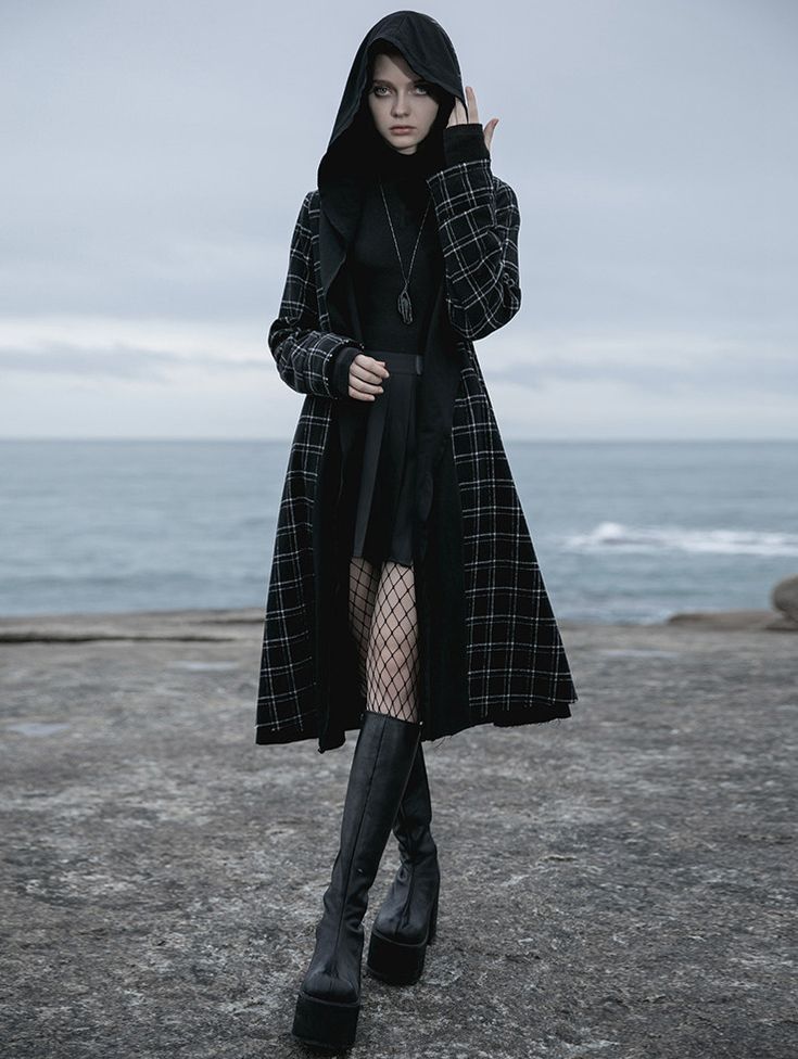 the image shows a young woman wearing a stylish black and white plaid hooded coat. the coat is long, reaching her knees, and features a large hood. she's wearing a black turtleneck top underneath, and a black pleated mini skirt. her accessories include fishnet stockings and high platform boots, both also in black.  the model poses outdoors, on a rocky beach near the ocean, which adds a dramatic backdrop to her gothic fashion. the overall aesthetic is dark, moody, and stylish, evoking a gothic or dark academia feel.