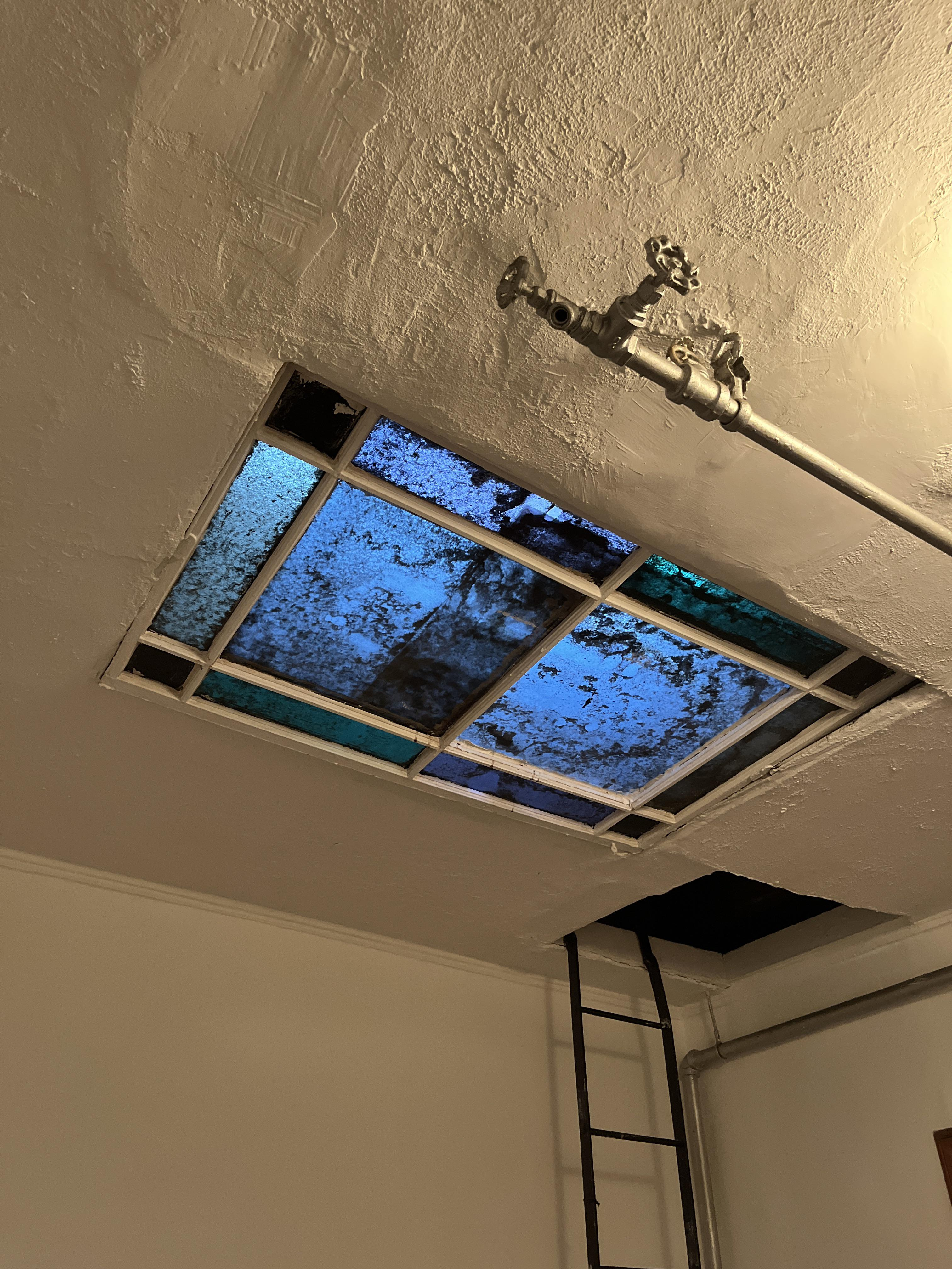 a lowangle shot of an indoor space showcasing a rectangular stainedglass skylight embedded within a textured white ceiling. the skylight, composed of blue and greenishblue glass panes, reveals a glimpse of a dim, cloudy sky. a white water pipe runs horizontally across the ceiling, its valve prominent. to the right, a black metal ladder suggests access to another level, perhaps an attic or loft. the lighting appears to be artificial and subdued.  the overall impression is one of an industrial or converted space with unique architectural features.