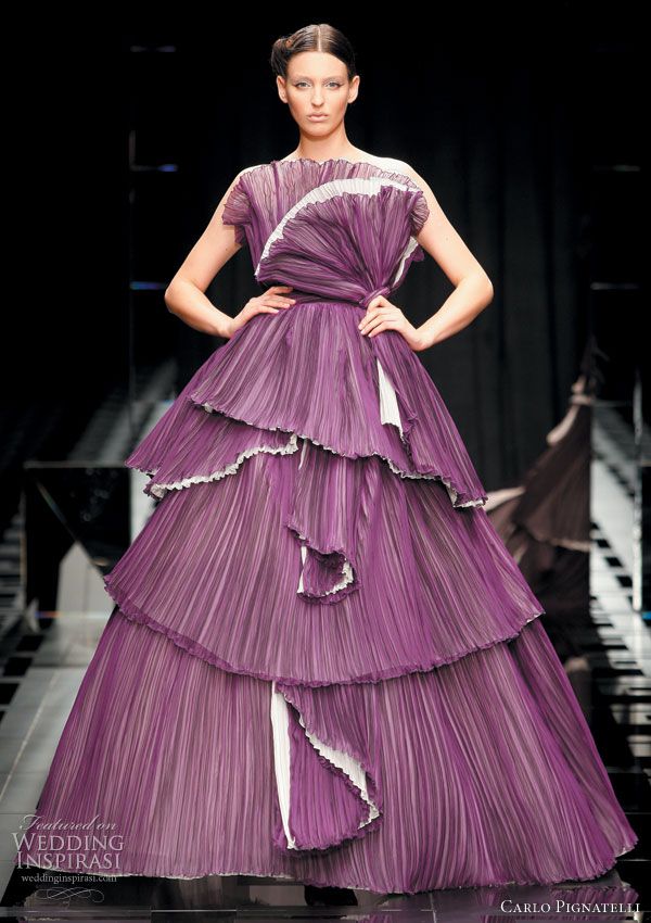 a model wearing a purple layered pleated dress with white ruffle detailing walks the runway. the dress is elegant and glamorous with a dramatic silhouette. the model is wearing her hair pulled back from her face and a minimalist makeup look. she is striking a pose for the photographers. the background is blurred, showcasing the dress as the focal point. the word "carlo pignatelli" appears in white at the bottom right of the image.