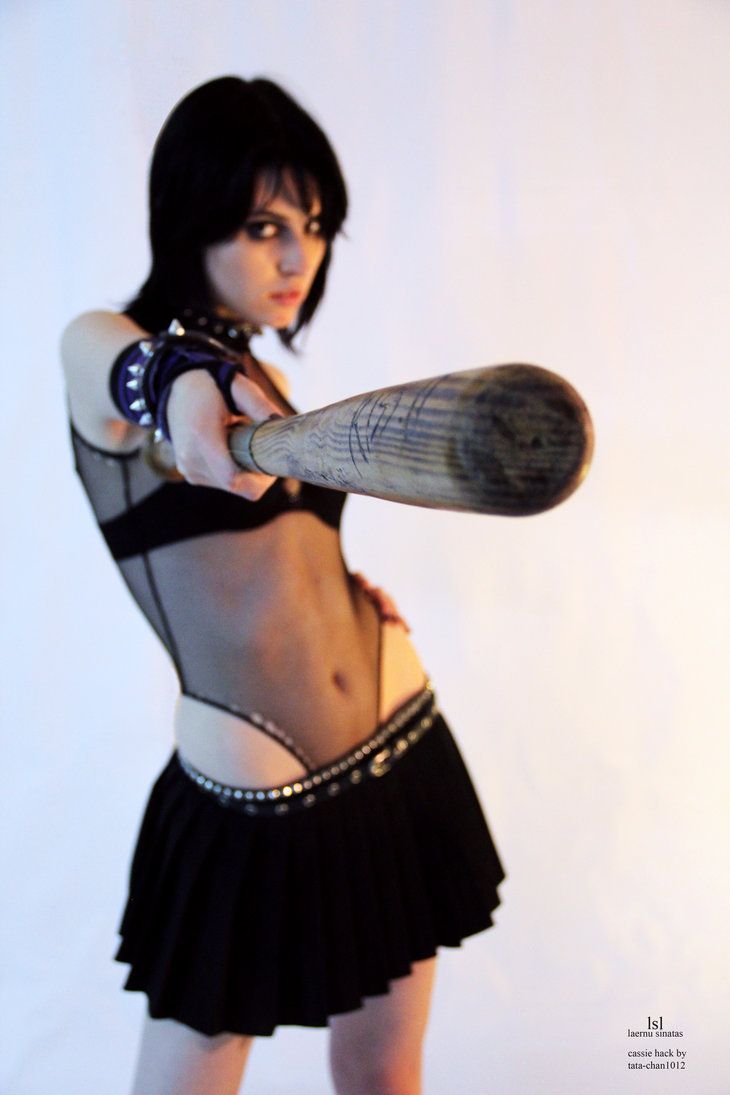 a young woman in a black mesh bodysuit, mini skirt and silver studded belt, with dark hair, is holding a wooden baseball bat in front of her face as she looks directly at the camera. the image is a portrait shot taken in a studio against a white background.