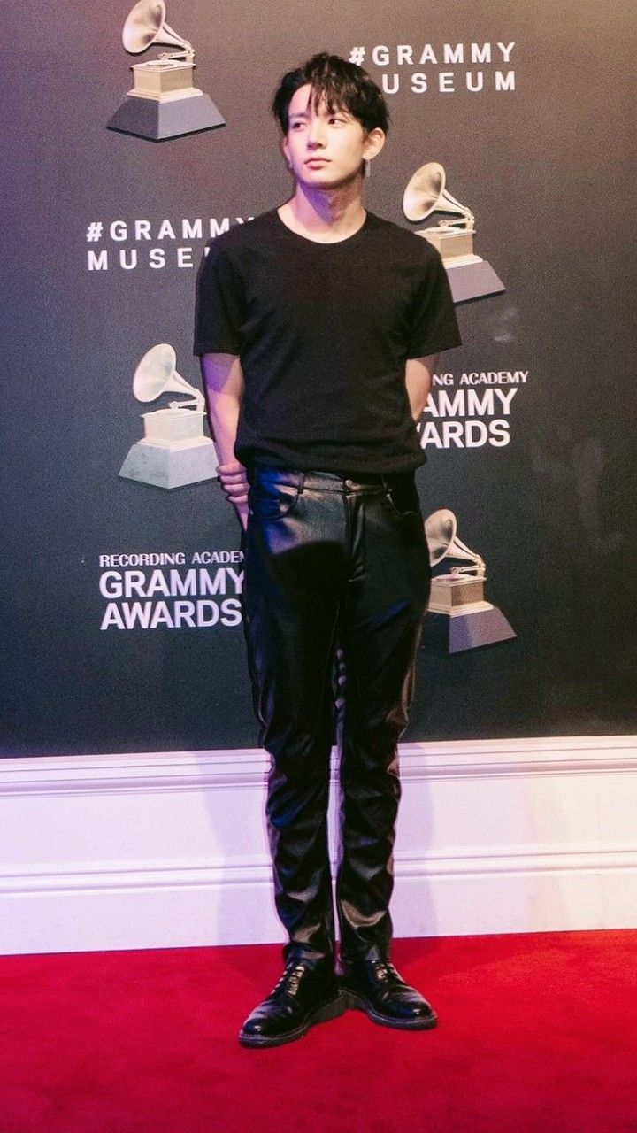 a full shot of a man standing on a red carpet in front of a grammy museum backdrop. he is wearing a black tshirt, black leather pants, and black shoes. he is looking off to the side.