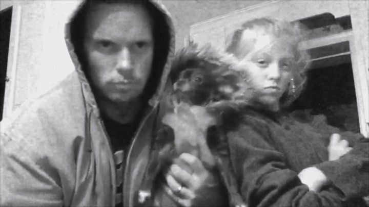 a black and white photo shows a man wearing a hooded jacket and a woman with their arms crossed, both with serious expressions. a small pet is positioned between them. the photo appears to be taken indoors.