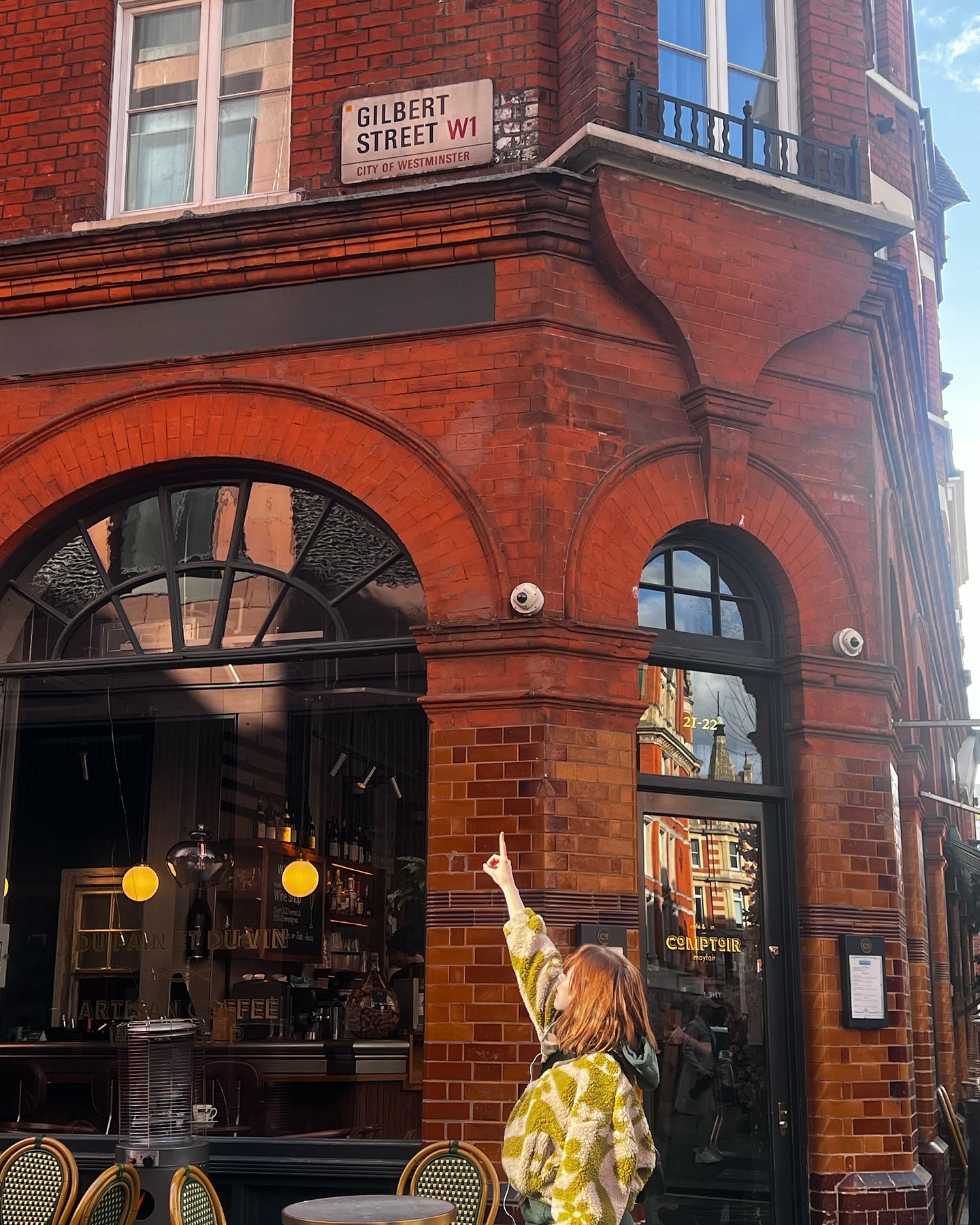 the image shows a woman standing on gilbert street in london's west end (w1), pointing up at the street sign. the street sign is affixed to a beautiful, old red brick building.  the building appears to house a pub or restaurant with large arched windows and outdoor seating visible. the woman is wearing a light green, textured sweater. the overall ambiance is that of a sunny autumn day in a vibrant london neighborhood.  the reflection in the windows shows other buildings and aspects of the surrounding street.