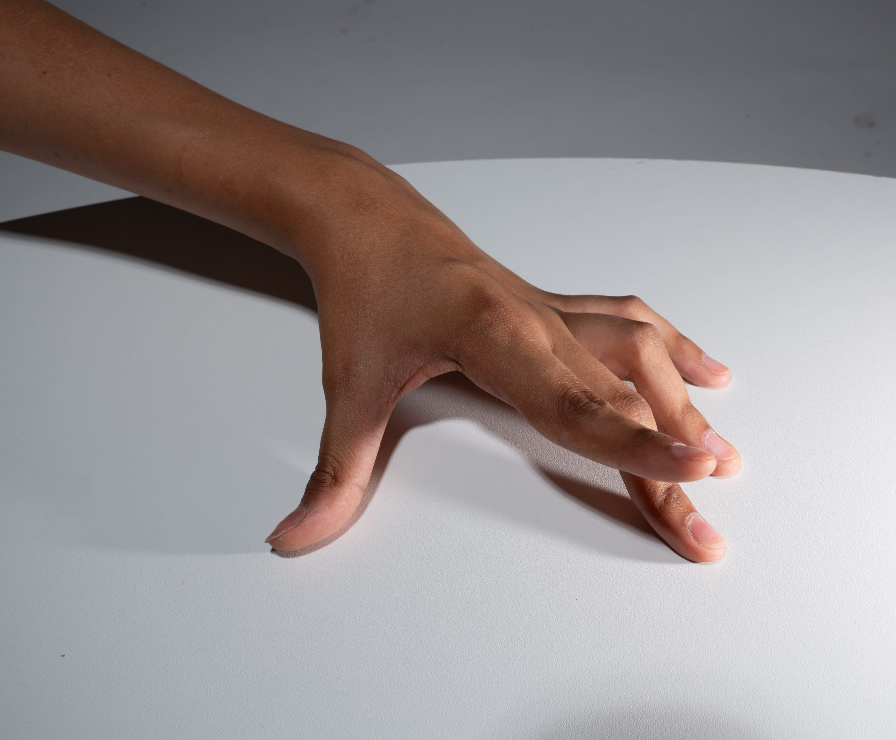 a hand with light brown skin is splayed open on a white surface. the hand is reaching forward as if to grab or touch something. the background is a light gray color. the image appears to be taken in a studio setting with artificial lighting.