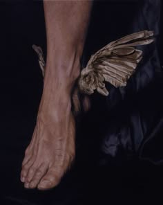 a close up of a human leg and foot. the leg is positioned so that the foot is pointing towards the viewer. the skin is flesh toned and appears to be nude. there is a brown, leathery, winglike object near the foot, possibly a feather. the background is dark.
