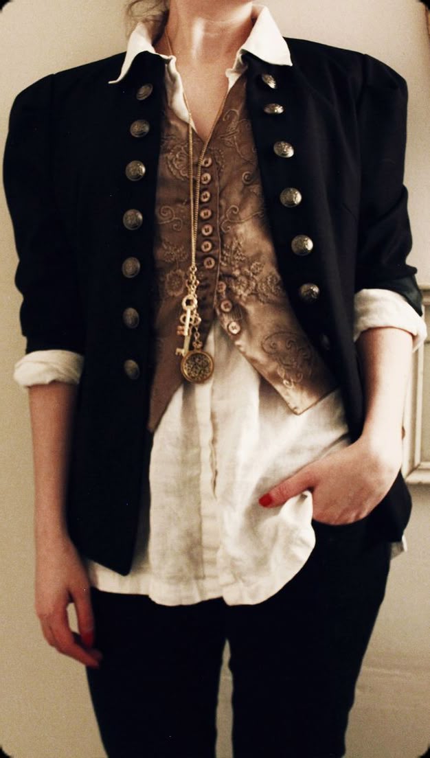 a woman is standing and posing for a photo. she is wearing a white collared shirt, a tan vest with buttons, a black jacket with large buttons, dark dress pants, and a long necklace that has a key and fob. she has red nail polish on and her right hand is in her pocket.
