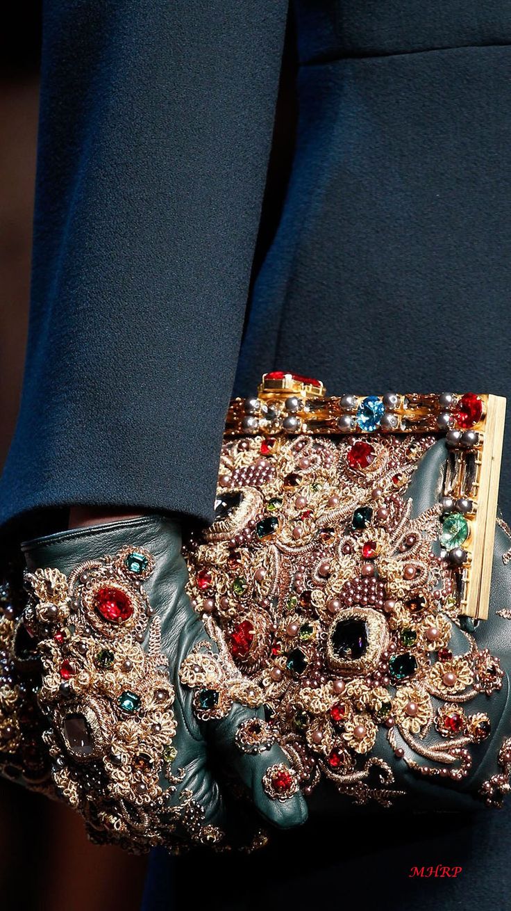 the image is a closeup detail shot showcasing a luxuriously embellished clutch and matching gloves.  the accessories are predominantly emerald green leather, heavily adorned with intricate gold embroidery, pearls, and various colored gemstones (including prominent red and other jewel tones).  the style is opulent and ornate, indicative of highend fashion. the gloves and clutch are displayed against a dark teal fabric, possibly a garment.  the overall feel is sophisticated and rich. the watermark "mjlrp" is visible in the bottom right corner.