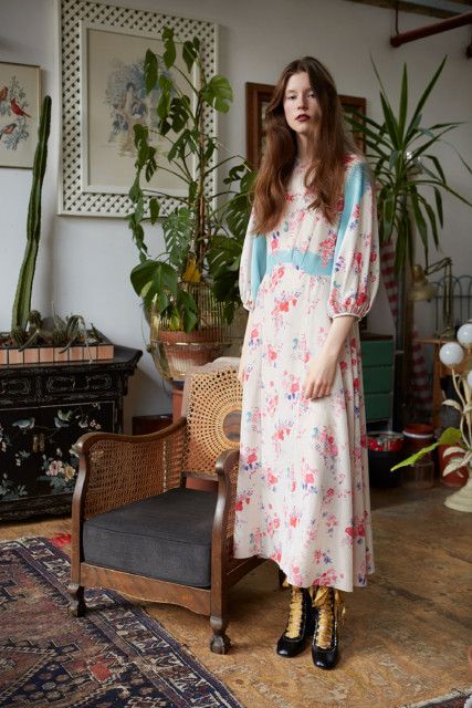 a fulllength shot of a woman in a vintagestyle, floral midi dress with lightcolored, long sleeves and a lightcolored bodice. the dress features a unique panel design in blue and white. the woman is wearing gold and black ankle boots. she is standing in a living room with plants, an antique wooden chair and a dark wooden cabinet. the room has an eclectic, vintage aesthetic. the overall feel of the image is a retro or vintage fashion shoot.
