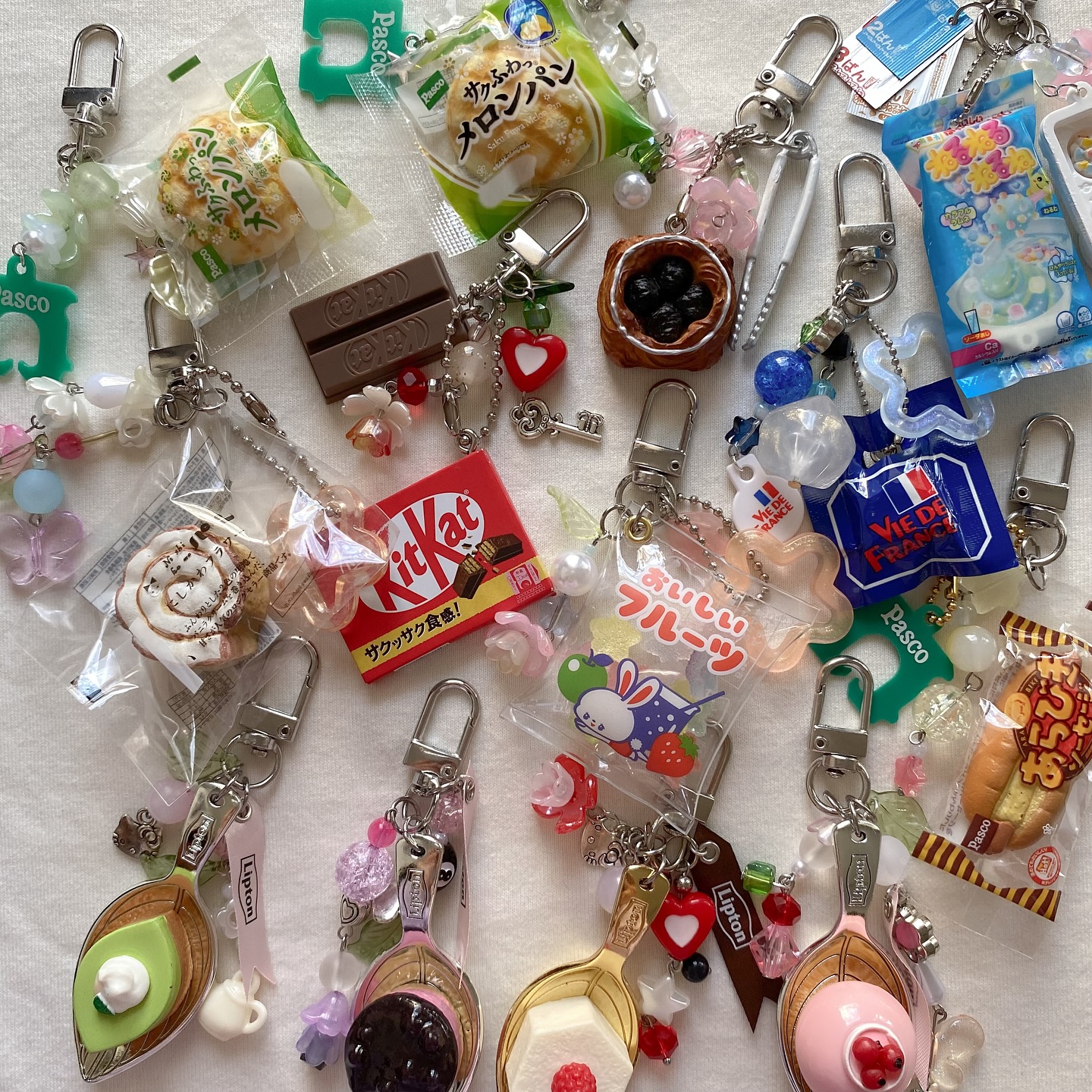 an overhead shot reveals a collection of keychains, each adorned with miniature food and pastry charms, along with other sweets and trinkets. keychains feature miniatures of pastries, a kitkat bar, melon pan, pasco products, fruit themed items, and lipton tea themed charms, all displayed on a white background.