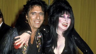 a portrait of a man and a woman. the man is on the left wearing a leather jacket with a black shirt underneath and the woman is on the right wearing a black dress. the woman is known for her gothlike style. they are both smiling at the camera and they have a romantic pose together.