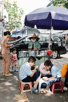 the image shows a bustling street scene with a woman serving food from a street stall. two young children sit on stools, eagerly eating their meal. a woman, possibly their mother, stands nearby, watching over them. an umbrella provides shade from the sun, and a car is parked behind the stall. the overall feel of the image is one of everyday life in a busy city.