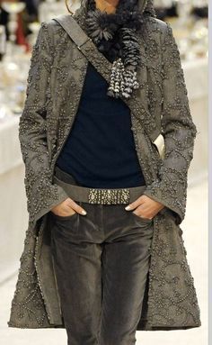 a woman is wearing a stylish grey coat, brown jeans, and a black shirt. she has on a fur scarf and a long, black necklace. she's posing with her hands in her pockets.