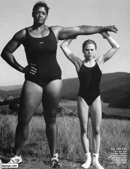 the black and white photograph showcases two women in swimsuits, standing in a grassy field.  the taller woman is significantly larger and more muscular than the smaller woman.  they are posed with the taller woman's arm draped over the smaller woman's shoulders, highlighting the dramatic size difference. the background shows a calm body of water and rolling hills. the image has a powerful aesthetic, emphasizing the contrasting body types and the strength of both athletes.  text in the lower right corner refers to the women by name and notes their contrasting sizes as being perfect for their respective sports.