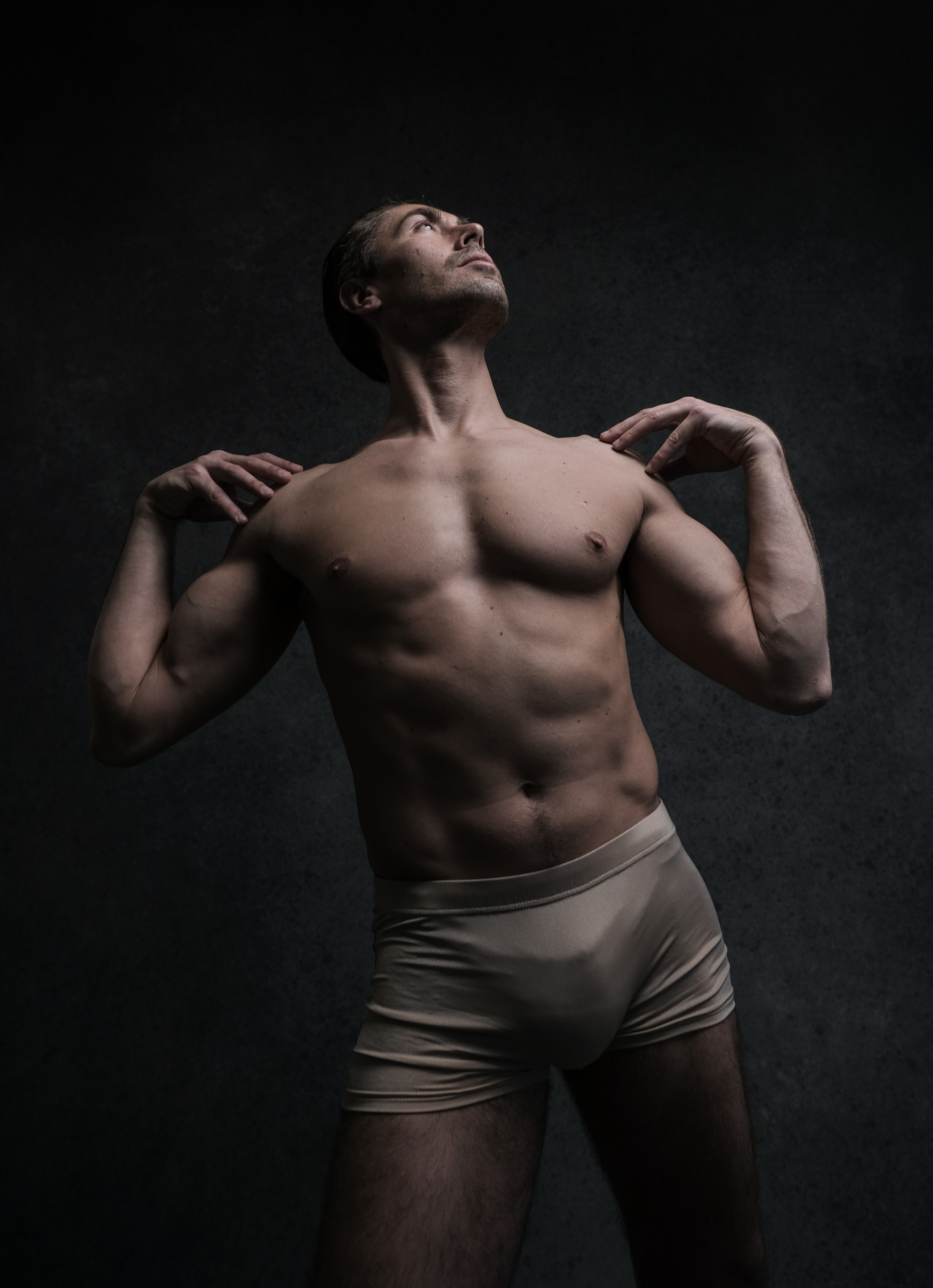 the image shows a muscular man standing in underwear in a dark room, with his head tilted back and eyes closed. the soft lighting highlights his muscular physique.  he has his hands resting on his shoulders. the image evokes a sense of strength and confidence, with an artistic aesthetic.