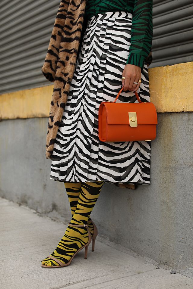 a fashionably dressed woman leans against a wall. she's wearing a zebraprint pleated skirt, a tigerprint fur coat, a green top, yellow tigerstriped socks, and nude heels. she carries a bright orange handbag. the setting appears to be outdoors on a city street.