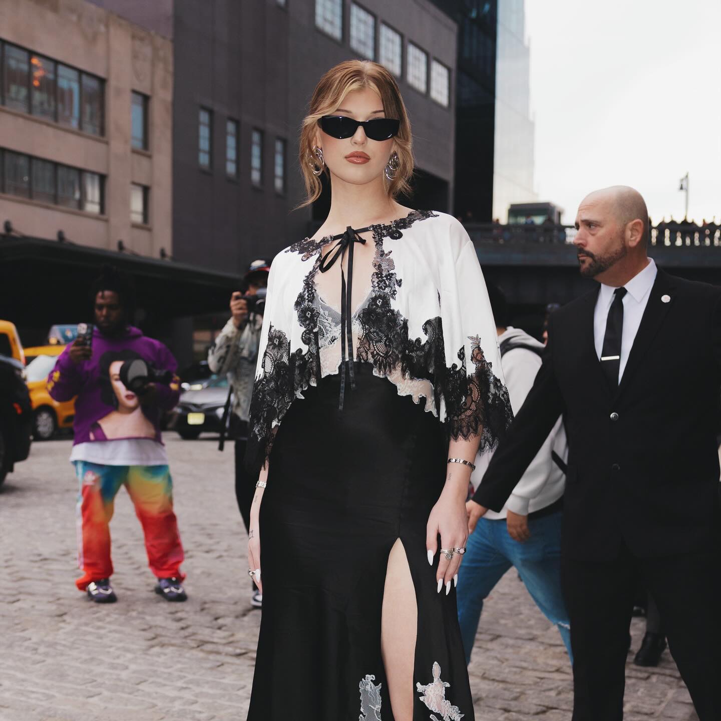 a woman in a black dress and white top with lace trim poses on a cobblestone street, flanked by a bodyguard in a suit. a photographer captures the scene, while other people walk by in the background. the setting appears to be an urban area with buildings and a yellow taxi visible.