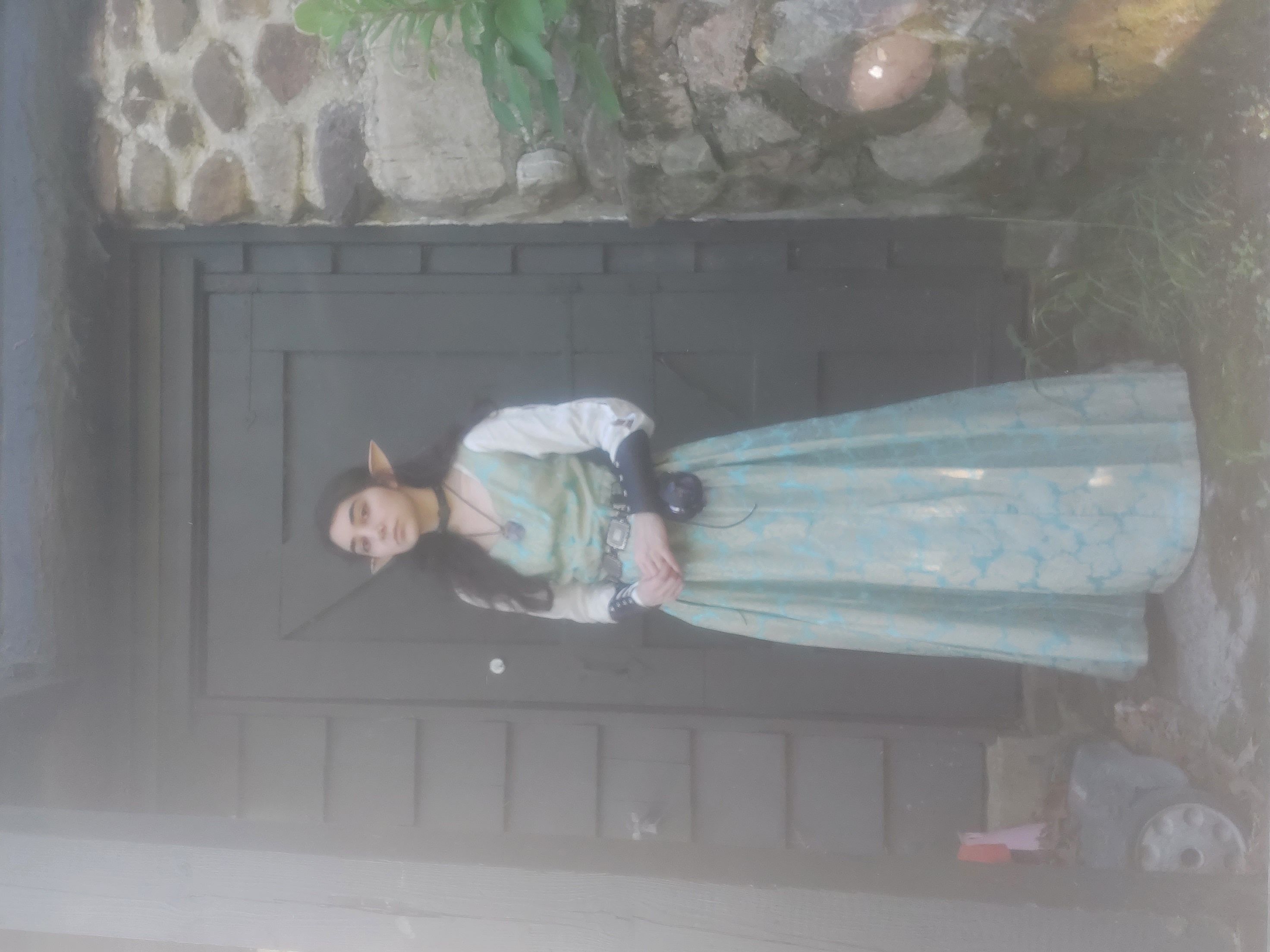 a young woman, dressed in an elaborate teal elf costume, stands calmly before a dark gray door set into a stone wall. the scene is softly lit, giving a misty or foggy atmosphere. her costume includes a long, flowing dress, a dark belt, and pointed elf ears.  the wall behind her is made of rough, uneven stones, some covered with moss. the overall mood is mysterious and evocative of fantasy or medieval settings.