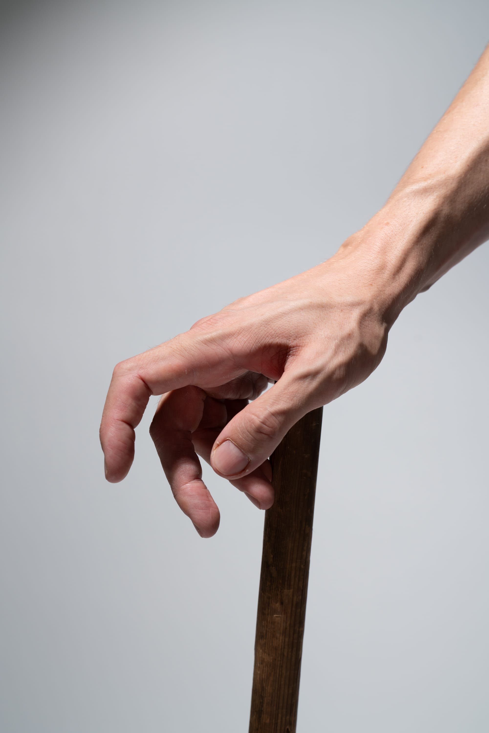 a closeup shot of a man's hand tightly gripping a wooden walking stick against a light gray background. the image emphasizes the strength and detail of the hand, including prominent veins and tendons, suggesting the stick's role in providing support and assistance.