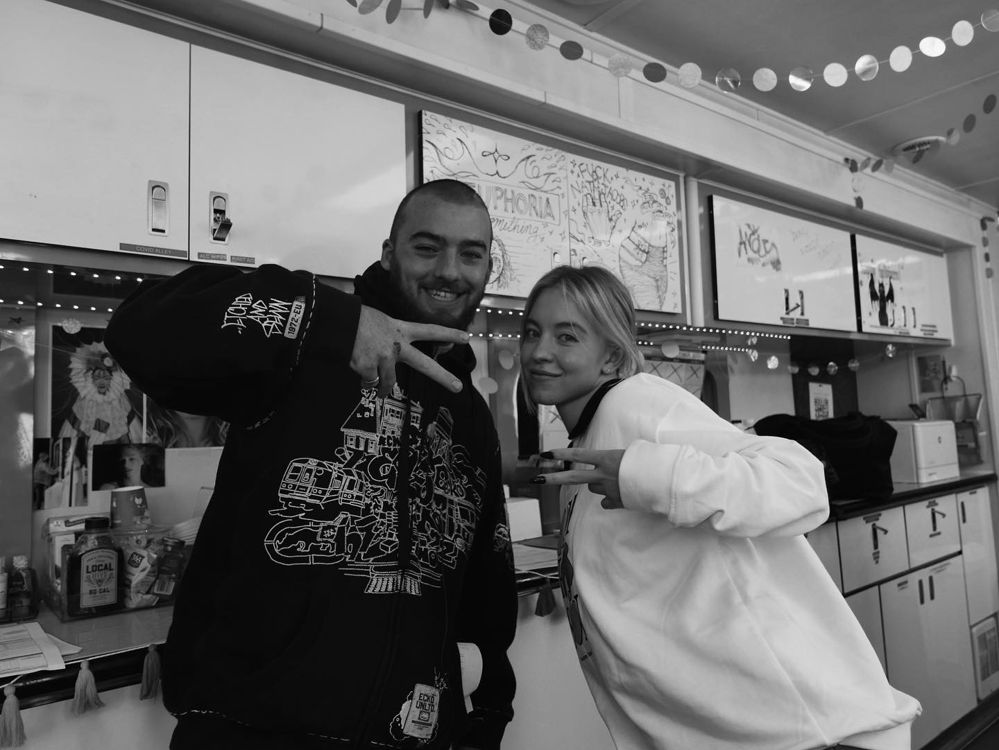 a black and white candid photo shows two young adults, a man and a woman, posing together with peace signs.  the man is wearing a black hoodie with white graffitistyle artwork, and the woman is wearing a white sweatshirt.  they appear happy and relaxed. the background indicates they are in a mobile recording studio or similar space with whiteboards displaying writing and drawings; the overall scene is casual and indicates a potentially artistic or musical environment.
