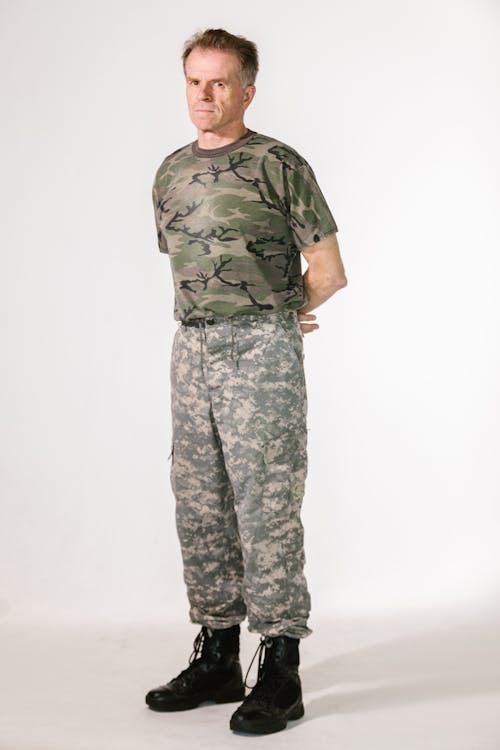 a fullbody studio shot of a man in military attire. he is standing with his arms crossed, looking directly at the camera. he is wearing a camouflage tshirt and matching camouflage pants. his expression is serious. he is wearing black military boots. the background is a plain white.