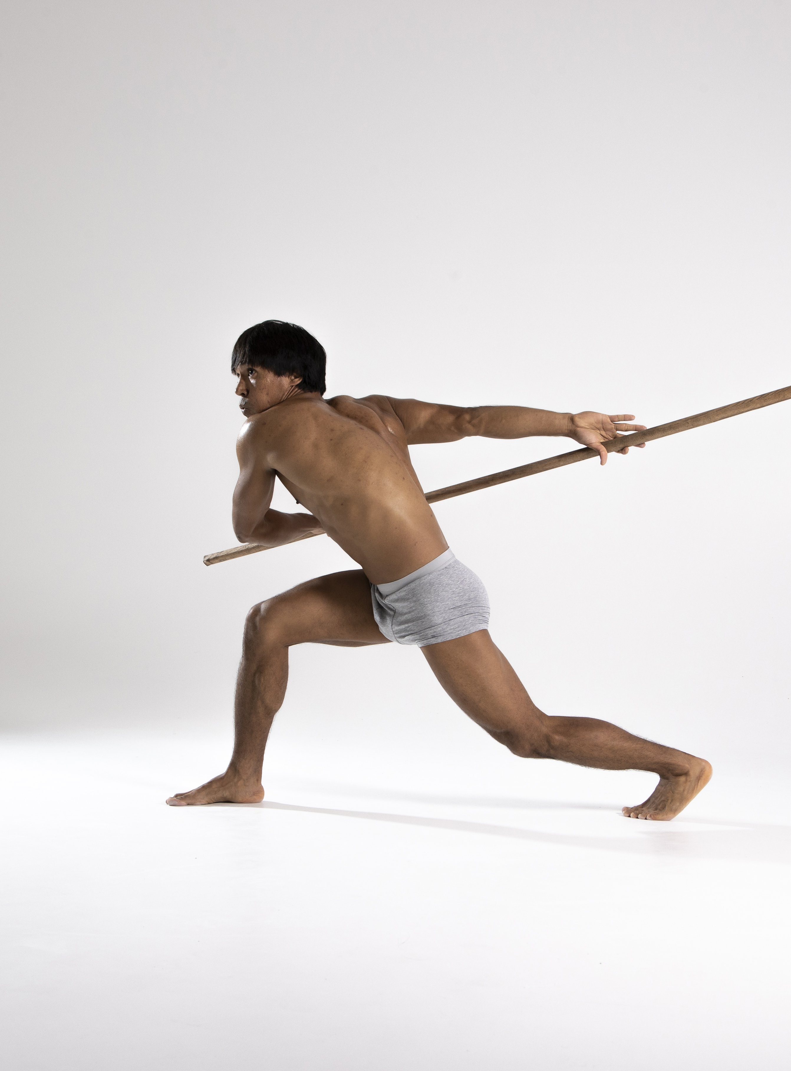 a shirtless man in a loincloth is lunging forward while wielding a wooden spear in a studio shot against a light background. he is barefoot and in an action pose, turning his upper body while holding the spear. his physique is athletic and he has indoaryan features.