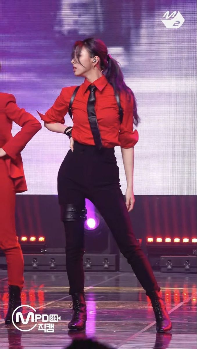 a female kpop singer performs on stage in a red shirt, black pants, and boots. she has her hair styled in a ponytail, and has a microphone in her hand. she is looking straight ahead with a determined expression. she appears to be in the middle of a performance, as the stage is lit with colorful lights.  the performance is being filmed by a camera,  and there is an mnet logo and "mpd" on the floor.