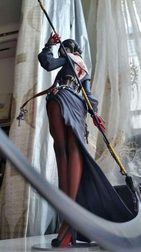 the image shows a figurine of a woman in an anime style holding a scythe. she is wearing a black dress with a high slit, exposing her legs, and black high heels with red soles. the figure has long hair and is wearing gloves. the costume includes a belt with intricate detailing and jewelry. the statue is standing on a clear base, and there are curtains in the background.