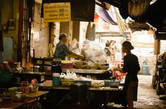the image showcases a bustling street food scene, likely in southeast asia. an older man in a blue shirt cooks at a wok, his station crowded with ingredients and supplies. steam rises from the wok, hinting at the delicious food being prepared.  a woman in a dark shirt stands nearby, perhaps waiting for her order. the scene is bathed in warm, natural light, creating a dynamic contrast of light and shadow. the overall atmosphere feels authentic and full of life.