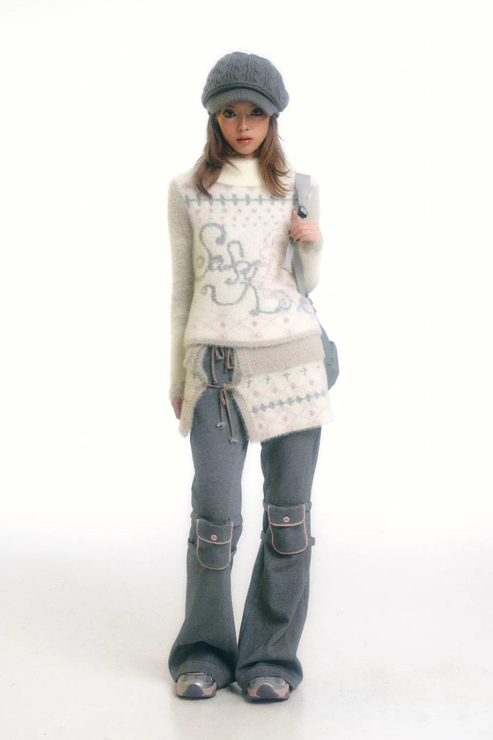 a fullbody shot of a young woman model posing in a studio against a white background. she is wearing a gray cable knit cap, a light beige fluffy sweater with a pattern, gray pants with small pockets at the knees, and gray shoes. she is holding a gray bag over her shoulder.